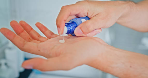 istock Young man smiling while applying moisturizer to his face in front of mirror in the bathroom at home. Healthy lifestyle in morning routine concept. 2200648242