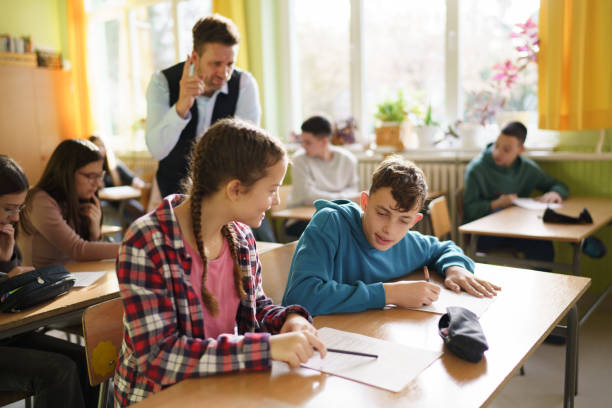 Concentrated Primary School Children Writing School Test on Geography or Ecology in Modern Classroom. Female Teacher Walking Between Desks, Controlling Group of Diverse Kids During the Exam Process.