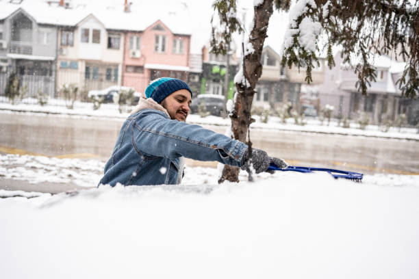 A man in warm clothing clears snow with a brush on a cold winter day, set against an urban residential area background.