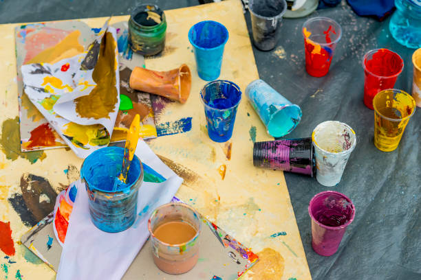 Cups filled with colorful paint scattered on a messy table with brushes and paper. Concept of creativity, art, artistic process