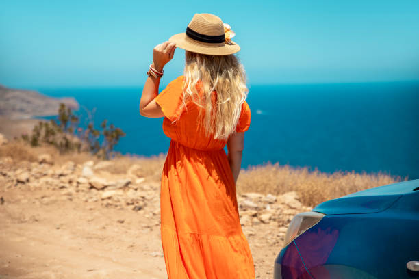 Unrecognizable Woman in orange dress with sun hat standing by the blue car and enjoying road trip during a Vacation in Greek island