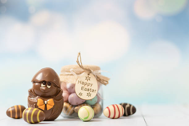 Chocolate easter chicken in shell and a glass jar with small chocolate easter eggs stands in the left on a white wooden table with little copy space on the right,close-up view from below.