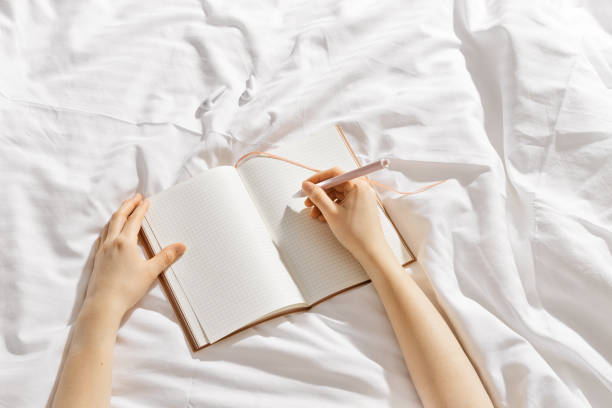 istock Woman writing on blank white page diary journal on white bedcloth in bed, holds paper notebook in hands, device free time before sleep or morning, digital detox, psychological wellbeing 2200719986