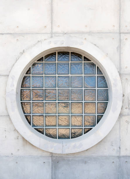 A circular glass block window embedded in a modern concrete wall. The window features a grid of textured glass bricks, allowing natural light to pass through while maintaining privacy, front view with the copy space.