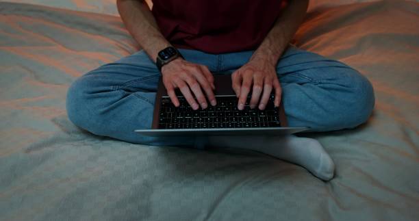 A man is typing on a laptop, standing on his feet, sitting in a lotus position on the bed, Close-up of male hands on the keyboard, no face, front view from above