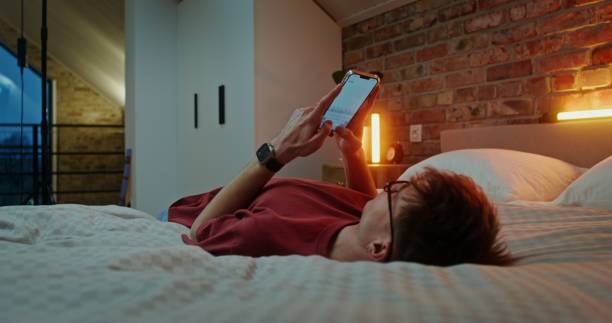istock Young man using smartphone while lying across the bed in stylish bedroom 2200756078
