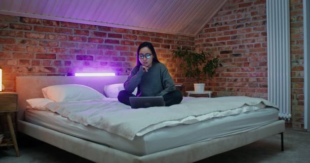 Young Asian woman in casual clothes smiling while video chatting on laptop, sitting on bed in stylish modern bedroom. The video moves from a long shot to a woman's face.