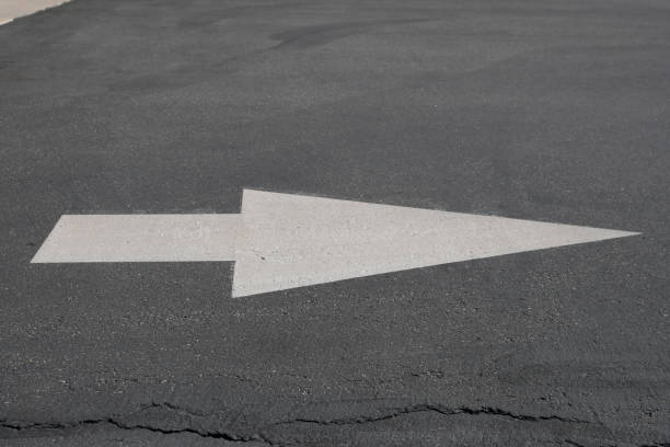 White arrow painted on dark asphalt, indicating direction. The arrow is large and prominent with a wide base and pointed tip, showing a simple, clear design. The pavement is slightly cracked and worn, suggesting outdoor exposure and regular use. This type of directional sign is commonly found in parking lots, roads, or driveways to guide vehicles. The texture of the asphalt contrasts with the smoothness of the paint, enhancing visibility.