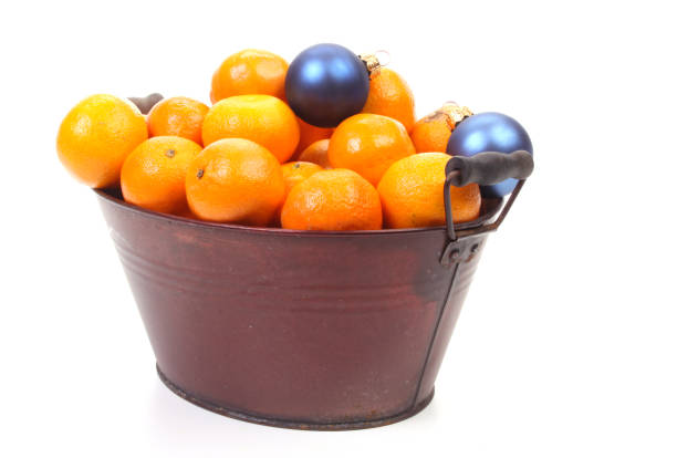 A close-up view of a holiday-themed still life arrangement on a rustic wooden surface, featuring vibrant orange slices from Citrus sinensis, cinnamon sticks from Cinnamomum verum, clusters of red cotoneaster berries, green pine branches from Pinus, and whole cloves from Syzygium aromaticum alongside black peppercorns from Piper nigrum. A white porcelain cup filled with steaming black tea sits centrally, accompanied by striped red-and-white candy canes, a woven basket, walnuts from Juglans regia, and scattered spices. Twinkling fairy lights in the blurred background add a soft glow, evoking a warm, cosy winter atmosphere indoors during the Christmas season. This image suits commercial applications such as holiday recipe illustrations, seasonal greeting cards, festive table setting features in lifestyle magazines, or editorial content on winter traditions for authors and media companies.