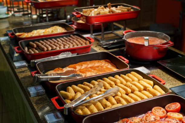 Close-up of a buffet display featuring baked chicken sausages, grilled tomatoes, crispy potato wedges, sausages, and creamy tomato sauce in vibrant red trays.