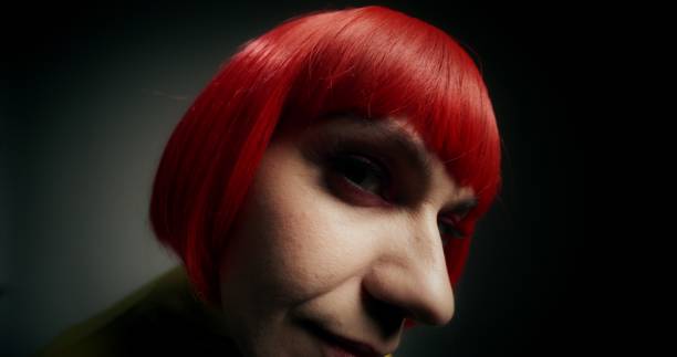 A woman in an orange wig poses approaching the camera and looking into it, standing on a gray background in the studio
