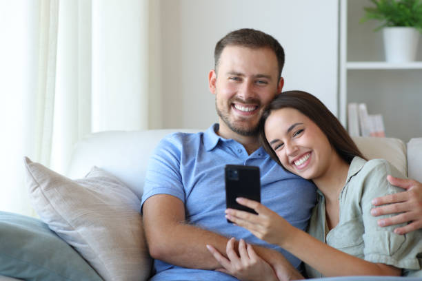 istock Happy couple holding phone looking at you at home 2200802208