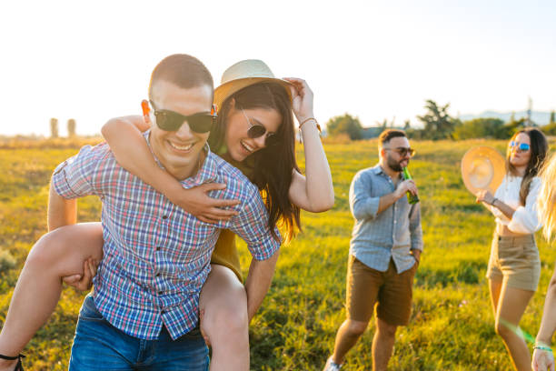 istock Young Man Giving His Girlfriend A Piggyback Ride While Their Friends Are Drinking Beer And Having A Dance Party In The Field 2200805510