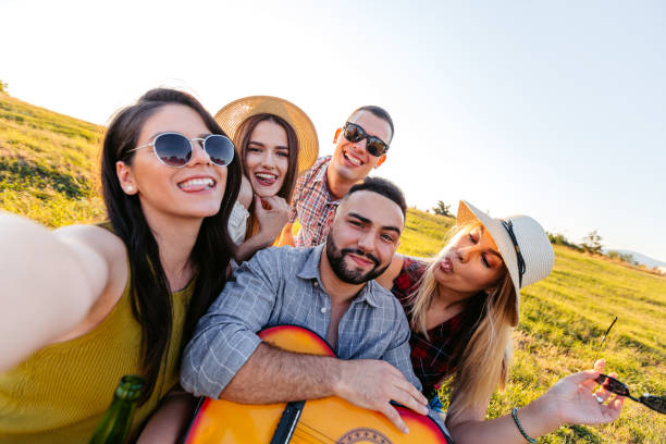 istock Group Of Friends Taking Selfies While Drinking Beer And Playing The Guitar In The Field At Sunset 2200806039