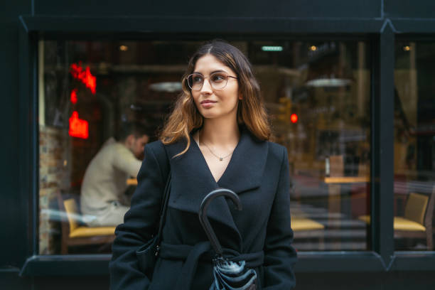 Confident young woman wearing glasses, standing beneath cafe window, gripping colorful umbrella while showcasing urban chic aesthetic Young adult woman with glasses holding an umbrella, standing in front of a cafe window, exuding confidence and style in an urban environment rainy cafe stock pictures, royalty-free photos & images