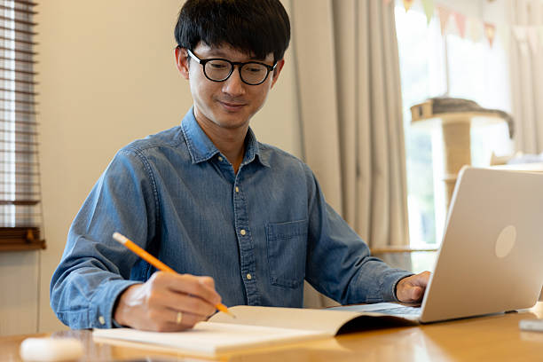 istock close up young asian man lecture on book while use laptop and open website to start study online bachelor degree course about foreign language or data analysis in home for people lifestyle and technology concept 2200828159