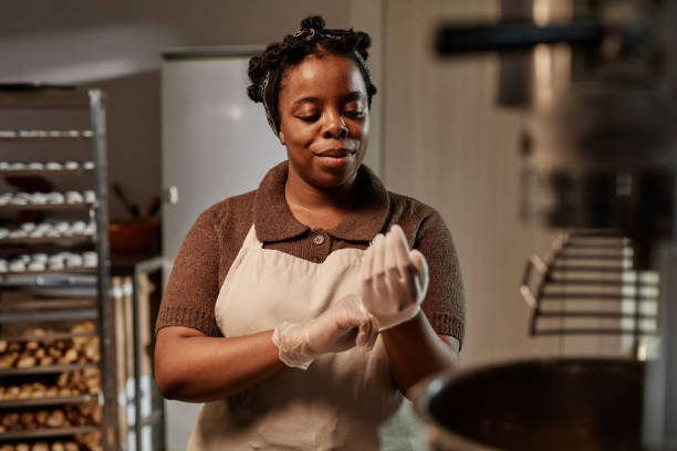 istock Smiling Black Female Baker Pulling on Vinyl Gloves Preparing to Cook in Bakery Kitchen 2200829680