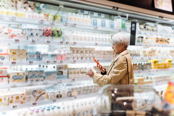 elegant asian senior woman choosing fresh milk and using smart phone to check the nutrition label on the pot.healthy family lifestyle. making healthier food choice. customer awareness concept. - dairy market hình ảnh sẵn có, bức ảnh & hình ảnh trả phí bản quyền một lần