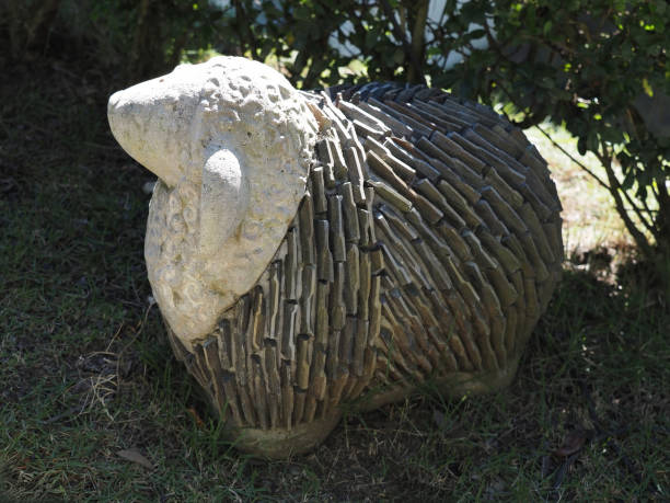 Decorative sheep sculpture made from stone sits in a garden surrounded by greenery in bright sunlight.