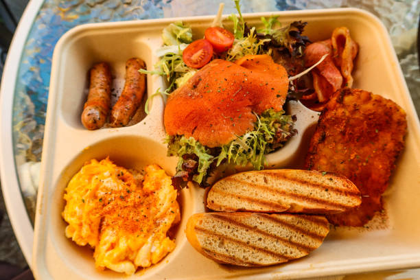 close up of a takeaway all day breakfast set in a paper box with divider. Smoked salmon, avocado, scrambled egg, toast, sourdough, sausages, vegetables salad, mushroom and bacons