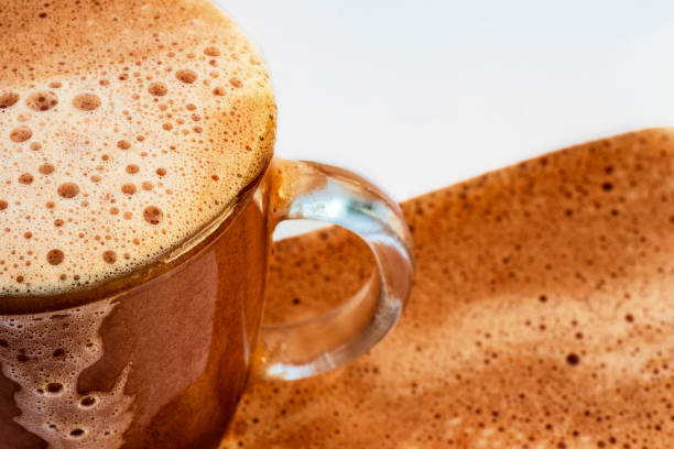 Part of hot foamy bubble chocolate beverage in glass mug. Drink overflows on mug and on white background, closeup.