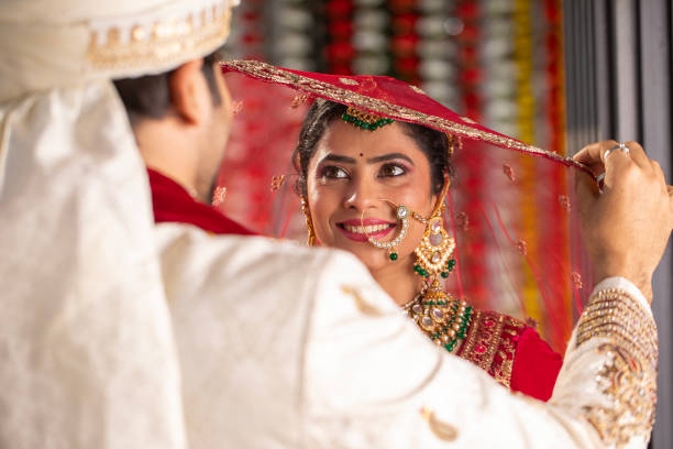 Beautiful bride in traditional clothing and jewelry smiling and looking at groom holding her red dupatta during wedding ceremony