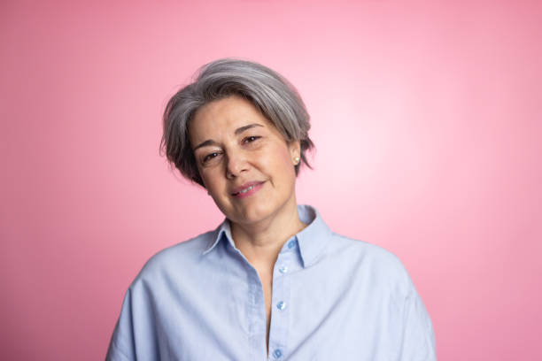 Studio portrait of a smiling mature woman with grey hair, wearing a light blue shirt, against a pink background