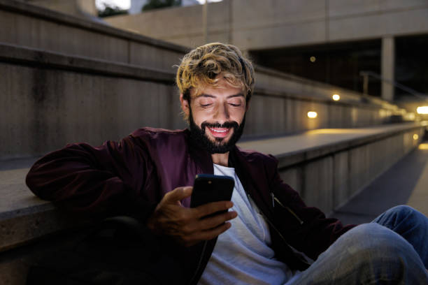 istock Smiling man using smartphone sitting on stairs at night 2200884531
