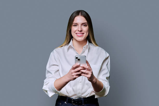 istock Young beautiful woman holding mobile phone, looking at camera on grey studio background 2200893968