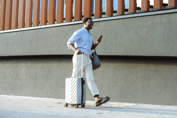 istock Man walking with suitcase near modern building while using smartphone 2200905035
