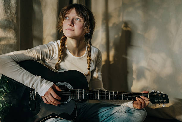istock Young woman playing acoustic guitar by window light 2200919464