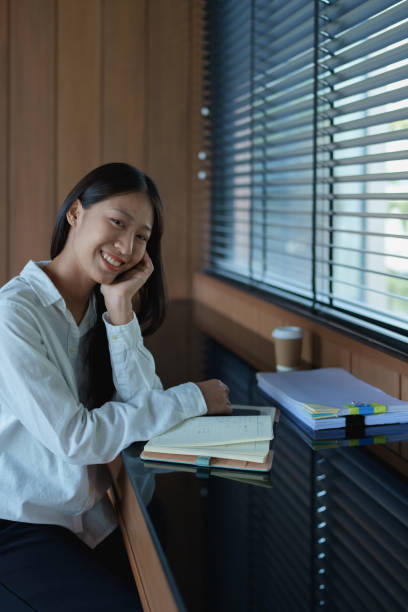 istock Young woman smiling while sitting at a desk with notepad, coffee, and natural light streaming through blinds 2200920627