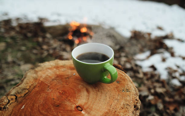 A cup of coffee on a stump and a fire surrounded by snow in the background. Winter camping concept.