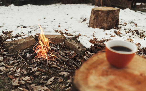 A cup of coffee on a stump and a fire surrounded by snow in the background. Winter camping concept.