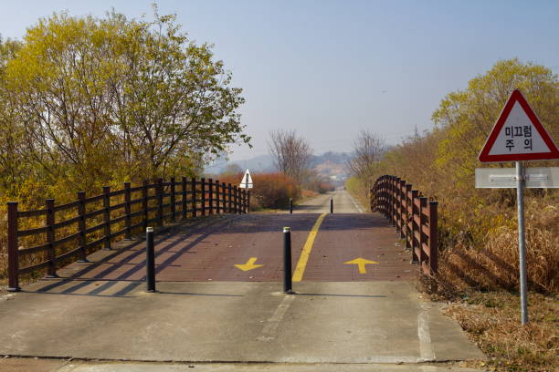 Cheongju, South Korea - November 11, 2020: A wooden bridge along the Ocheon Bicycle Path features a caution sign and safety barriers, guiding cyclists over a gentle incline amidst autumn foliage.