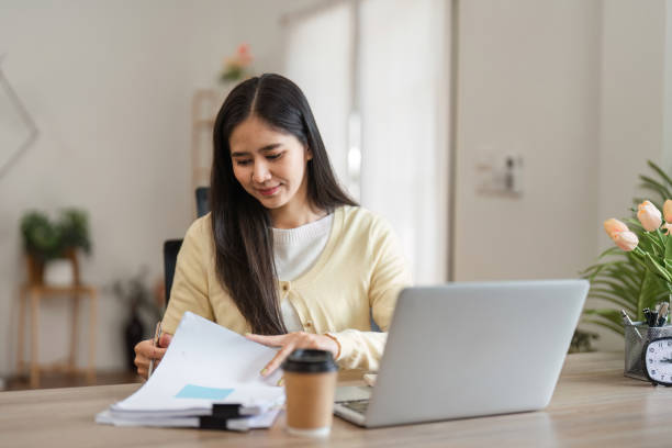 istock Concentrated young woman organizing papers while using a laptop in a cozy home office. 2200959748