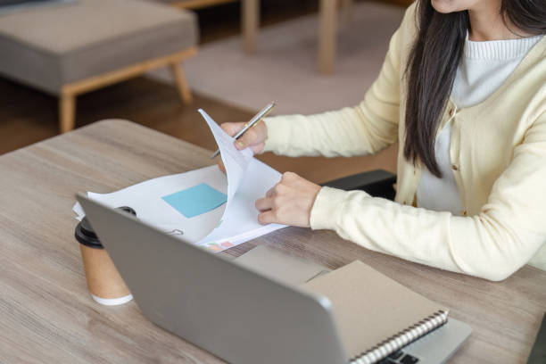 istock Focused Asian woman reviewing documents and taking notes while working on a laptop in a cozy home office. 2200959849