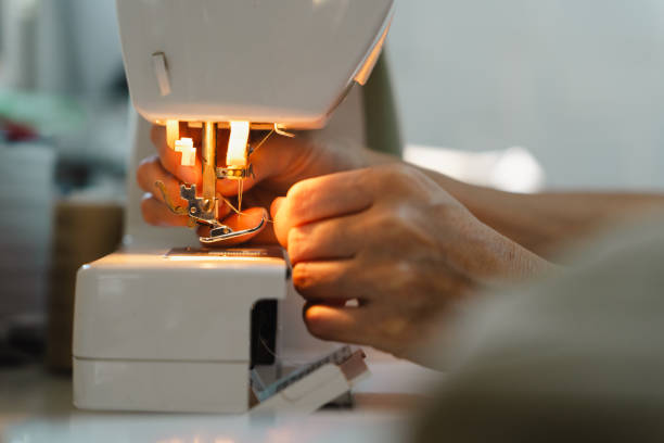 A person's hands are carefully threading a sewing machine. The light illuminates the machine's parts, highlighting the focus and precision of the task.