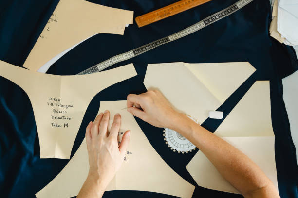 istock Top-table view of woman hands using a needle to arrenge craft paper patterns on fabric for sewing project, using measuring tape and tools on a dark fabric 2200961807