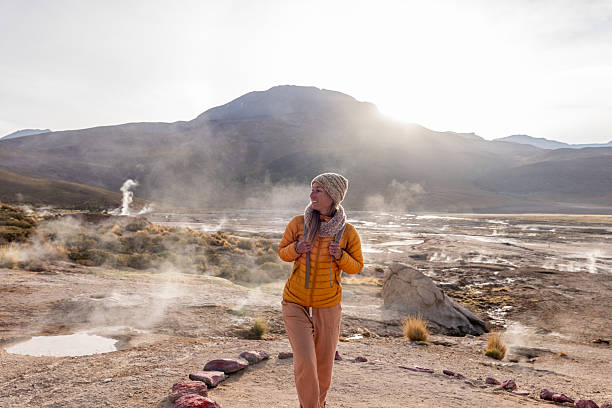 Smiling woman in yellow jacket and beanie exploring a steaming geothermal field. Enjoys outdoor adventure. Atacama desert, Chile