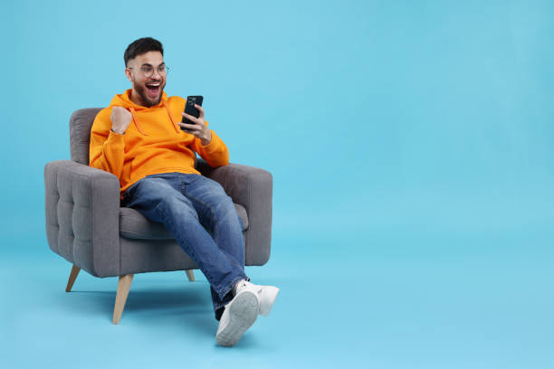 istock Happy young man using smartphone on armchair against light blue background, space for text 2200975271