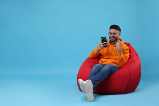 istock Happy young man using smartphone on bean bag chair against light blue background. Space for text 2200975290