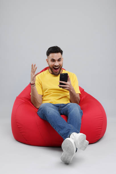 istock Emotional young man using smartphone on bean bag chair against grey background 2200975333