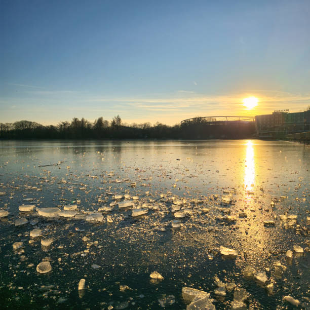 A picturesque and serene sunset slowly casting beautiful golden hues on sparkling icy waters, surrounded by lush trees Hannover Maschsee