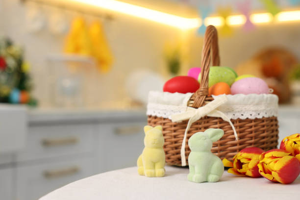 Easter decorations. Wicker basket with painted eggs, tulips and bunny figures on white table indoors, closeup. Space for text