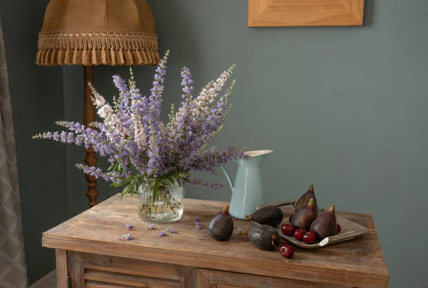 Rustic still life with fresh figs on a cork mat, a black plate, and a blurred background of vases and dried flowers, highlighting the figs' texture and color