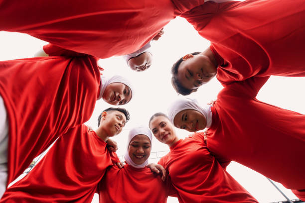 Low angle view of diverse group of young female football players, including hijabi athletes, huddling in a tight circle on the grass field before their match. Dressed in red jerseys and white athletic wear.
