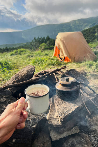 istock Hand holds white enamel mug with hot beverage next to rustic kettle on stone fire pit. 2200997467