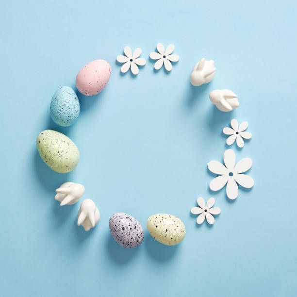 istock Minimalist Easter frame with white bunnies, colored eggs and wooden flowers on a blue background. Space for your text. 2201003373