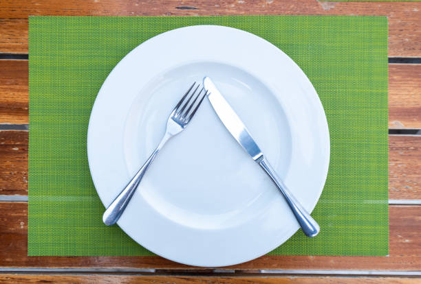istock a white ceramic plate with knife and fork arranged in a triangle shape on placemat atop a wooden table, stock image of Dining etiquettes background with spoon and fork arrangement 2201007731